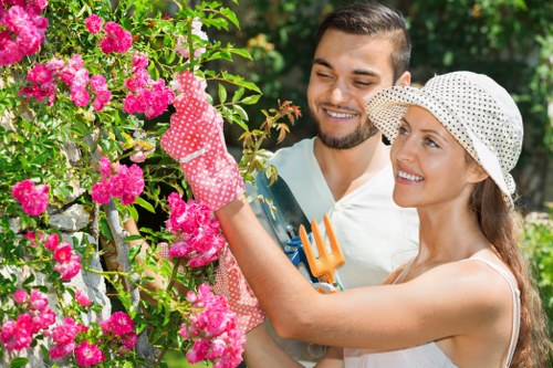 A smiling young couple is gardening outdoors in a lush, green garden, with the woman wearing a wide-brimmed hat and pink gardening gloves, holding a pruning tool, as she tends to a vibrant pink flowering shrub. The man stands close beside her, observing and supporting her work, with short dark hair and a neatly trimmed beard. The garden features well-maintained flower beds, dense foliage, and a variety of plants, suggesting a landscaped outdoor space suitable for gardening and lawn care services. Bright natural light illuminates the scene, indicating a clear, sunny day, with the couple engaging in a shared gardening activity typical of residential gardens in Croydon, with the backdrop of mature trees and climbing plants. This image exemplifies the outdoor environment managed by professional gardening services like Lawn Mowing Croydon, highlighting the importance of plant care, pruning, and outdoor beautification in maintaining a healthy and attractive garden in the local area.